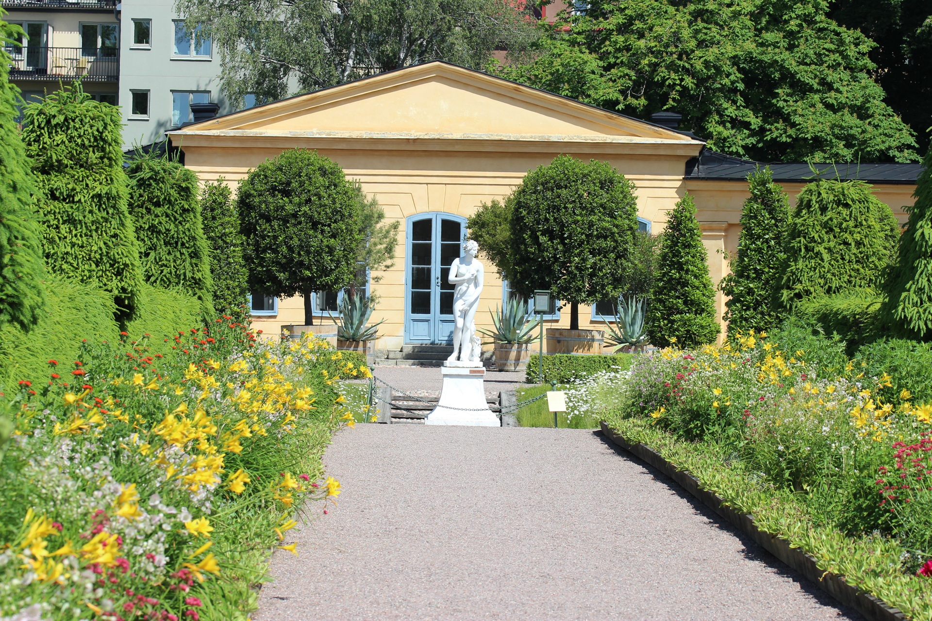 A graveled path bordered by red, yellow and white flowers. In the baakground a white Venus statue and a yellow stone building.
